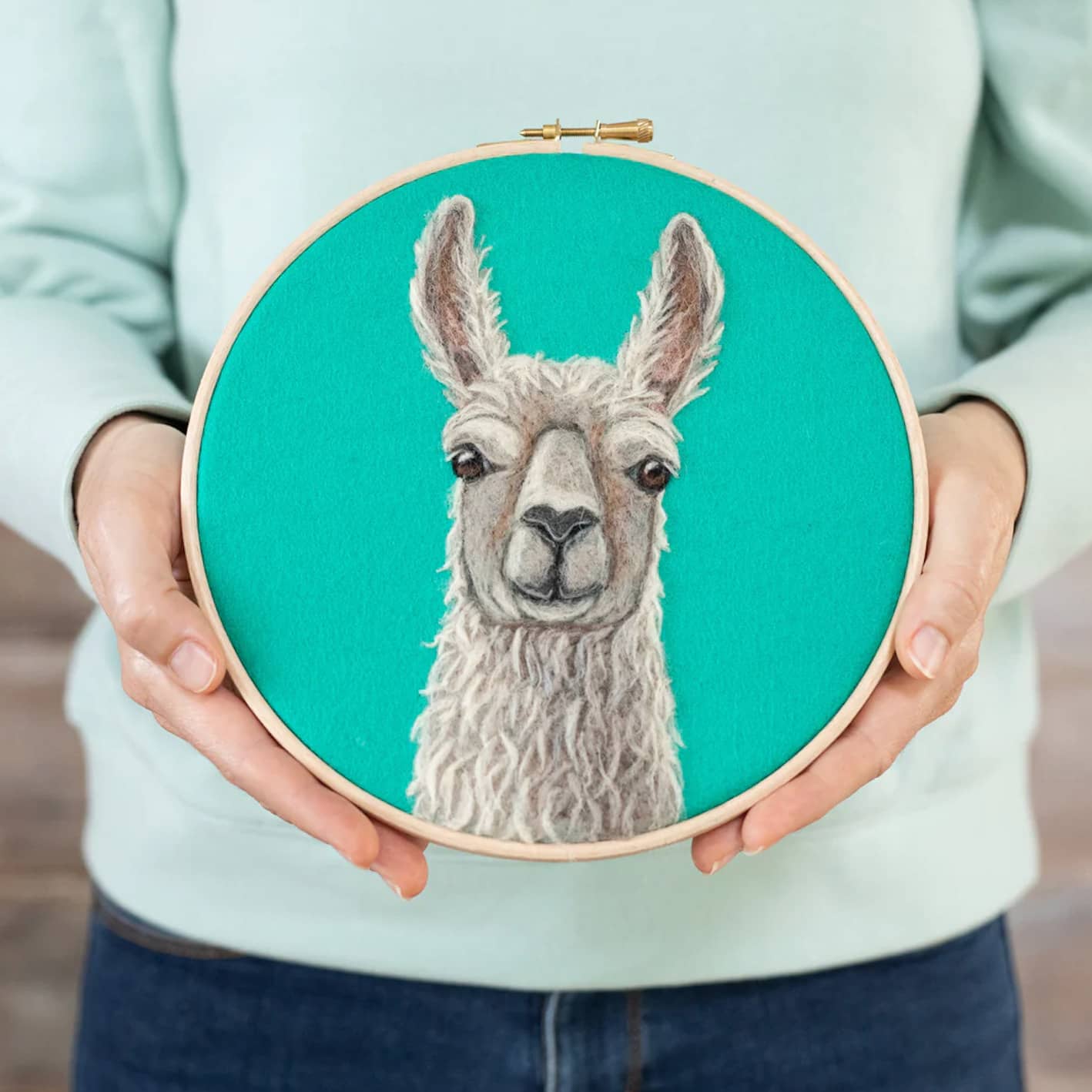 A woman is holding an embroidery hoop with a Llama Needle Felting Kit on it.