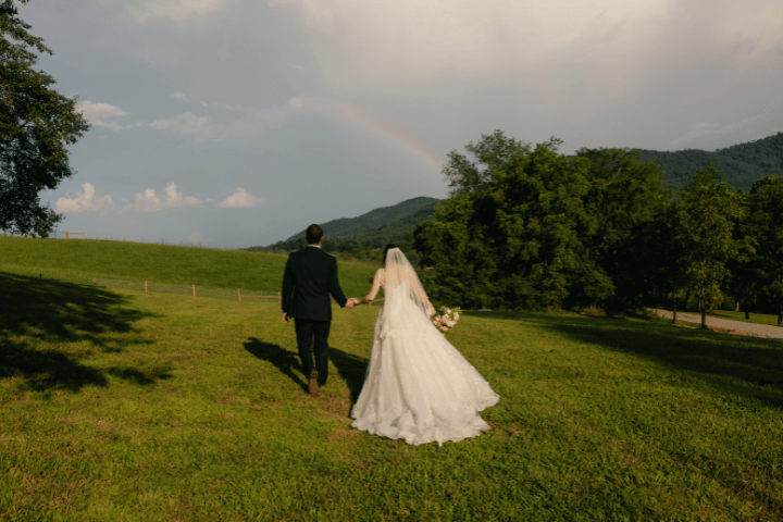A bride and groom stroll hand in hand through a green field, rainbow arching above, mountains and farm tranquility surrounding them.