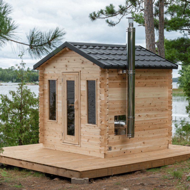 Cozy wooden cabin with a black metal roof and tall chimney rests on a deck among trees, overlooking the lake on a cloudy farm day.